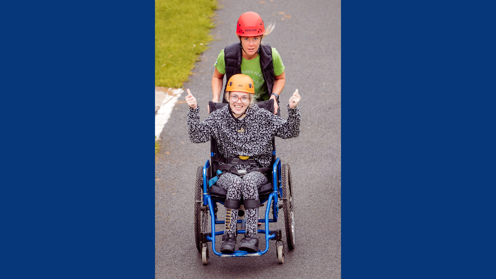 An image of a blonde girl in a blue wheelchair, wearing a hard hat and smiling at the camera giving a 'thumbs-up'. She is being pushed by woman in a green top and a red hard hat. 