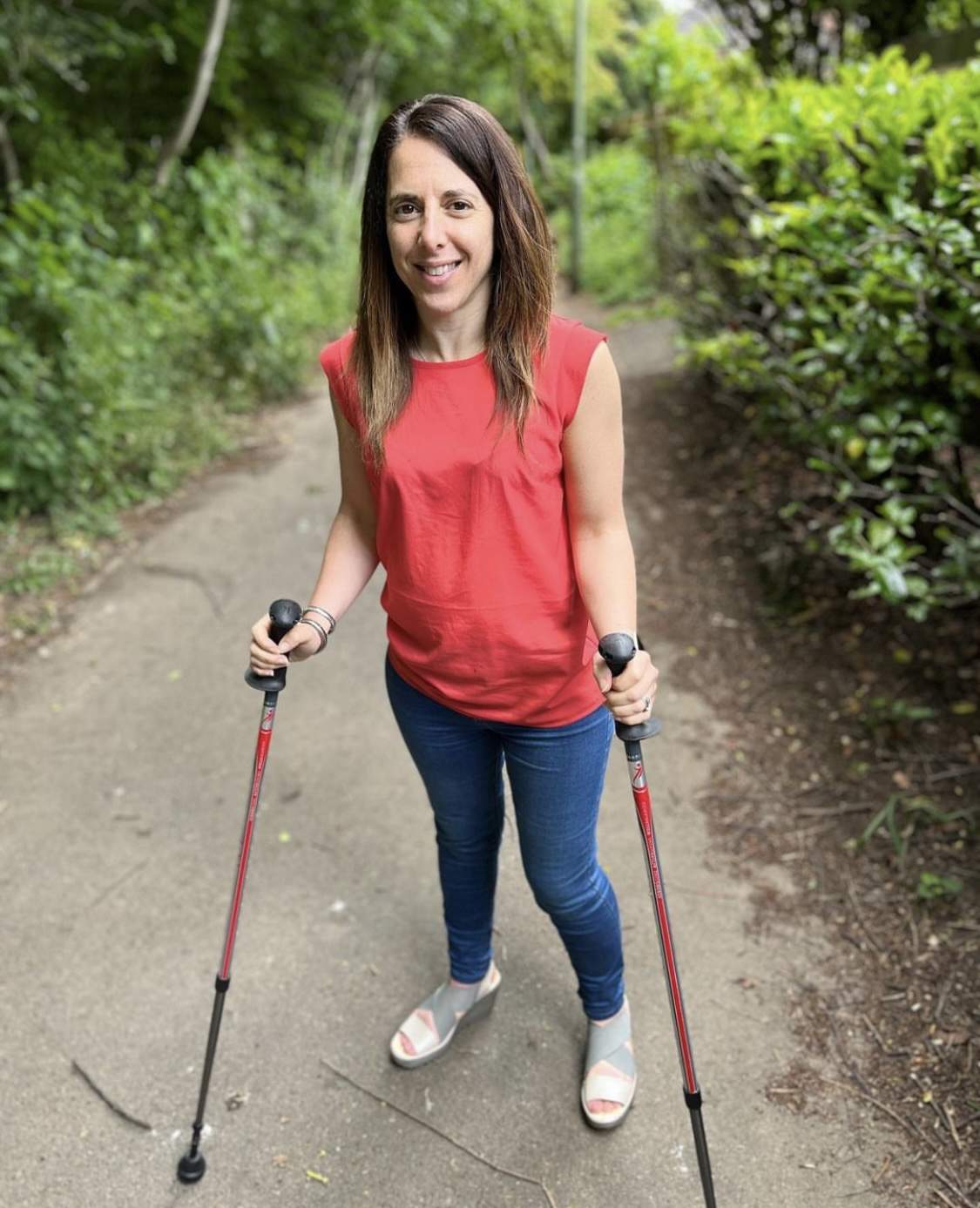 This image shows Georgina who is a woman standing on a nature path, smiling at the camera. She is holding two walking sticks for support. The woman is wearing a sleeveless red top, blue jeans, and light-coloured sandals. The path is surrounded by greenery, with trees and bushes lining both sides, creating a peaceful outdoor setting. The woman appears confident and relaxed as she walks along the path.