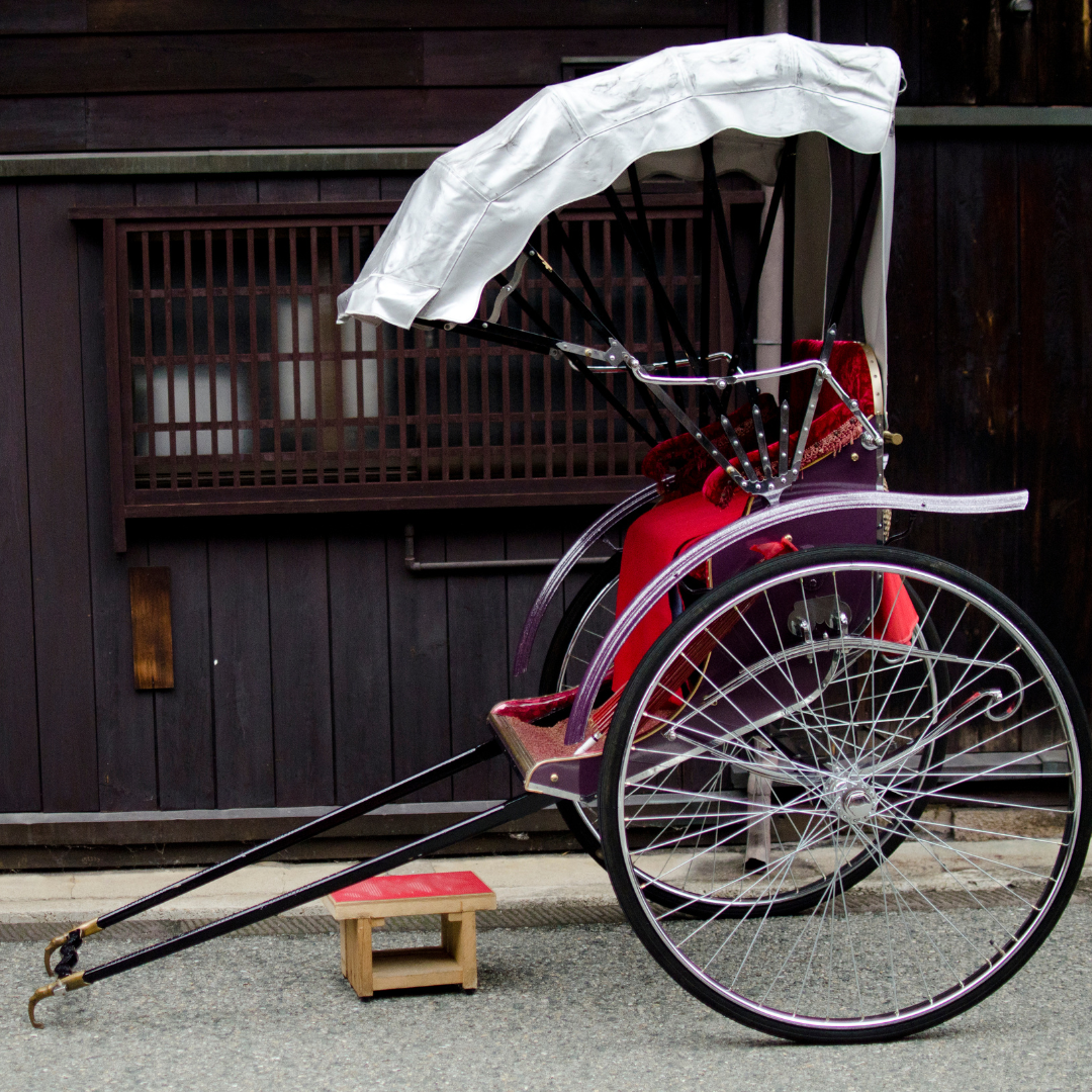 An image of a Rickshaw, which is a seat with two big wheels, and two handles at the front so it can be pulled along. There is a white cover, which acts as a roof.