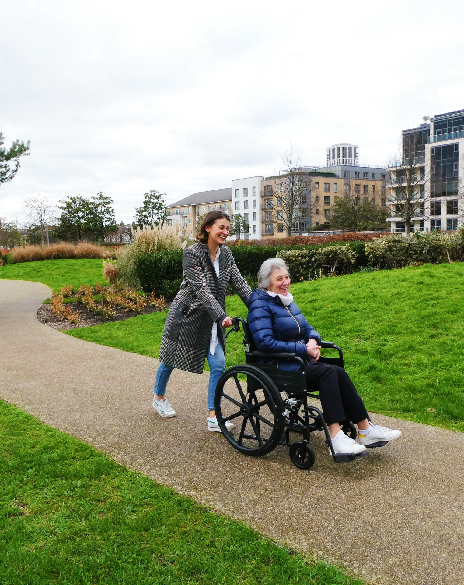 An image of a woman wearing a checked coat and blue jeans, smiling whilst pushing an older woman with grey hair and a navy puffer jacket in a wheelchair. 
