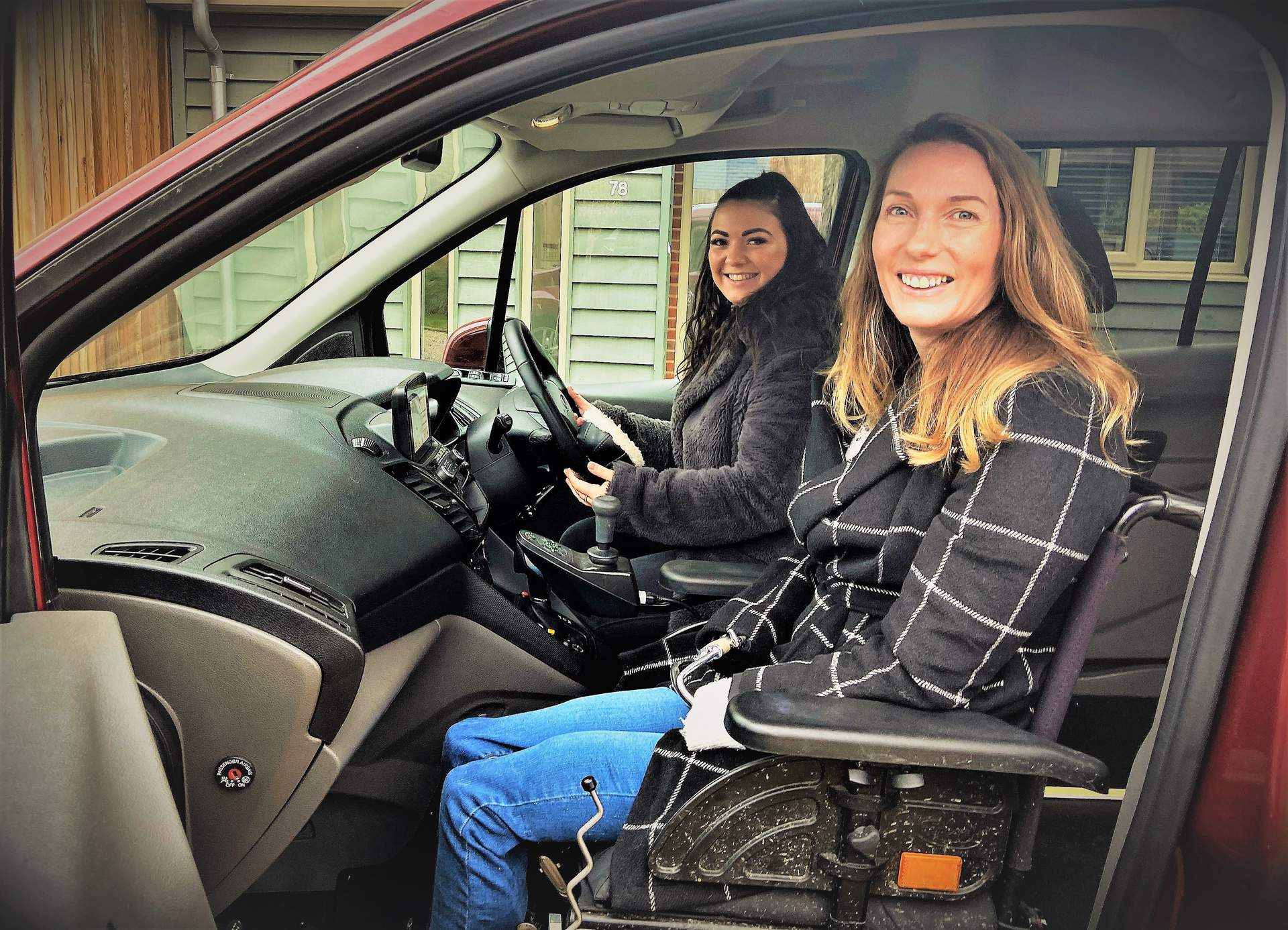 An image of a woman with light brown hair, sat in a wheelchair in the passenger seat space of the Ford Grand Tourneo Connect. There is another lady with dark brown hair, also smiling, sat behind the steering wheel of the car. 