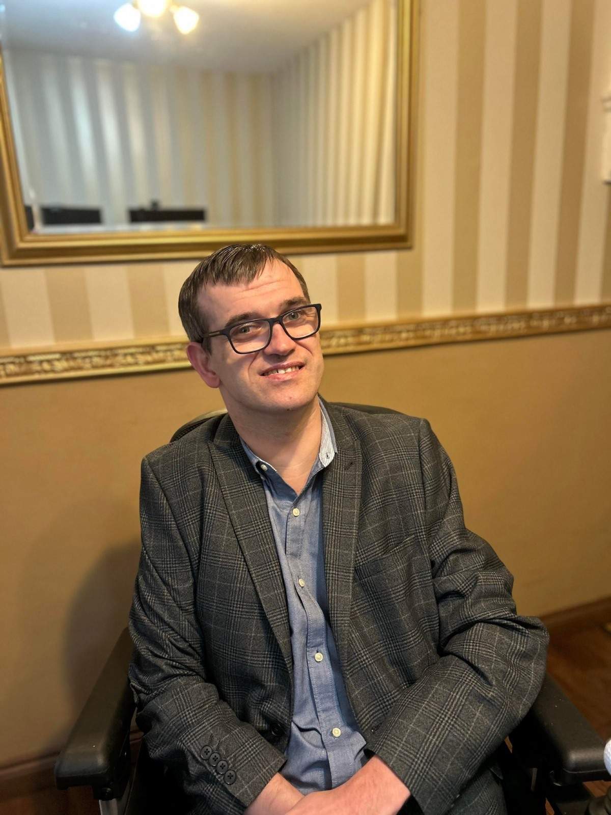  The image shows Lee, who is a man sitting in a chair and smiling at the camera. He is wearing glasses, a grey checkered blazer, and a light blue button-up shirt. His hands rest comfortably on his lap. Behind him is a wall with light-coloured vertical stripes and a gold-framed mirror reflecting part of the room's ceiling and lights. The atmosphere is warm, with soft lighting and a relaxed setting.