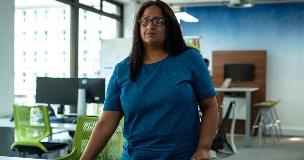 An image of an Asian woman with dark hair, black rimmed glasses, who is wearing a blue t-shirt. She has a neutral expression and is standing in an office space. There are green chairs and computers behind her. 