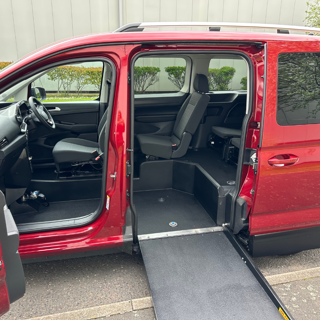 An image of a red coloured Ford Tourneo Connect which offers an upfront passenger option for the wheelchair user. There is a ramp to the side of the car which allows the wheelchair user to enter the car and be seated in the passenger seat.