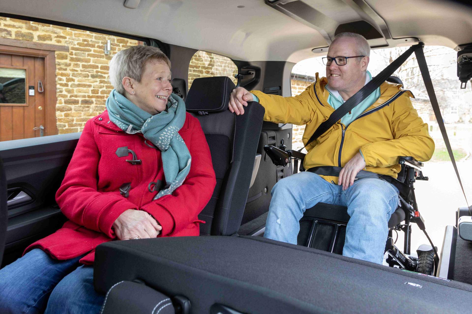 An image showing the rear passenger WAV of a man in a yellow coat and glasses, who is strapped inside the car in a wheelchair, with his seat belt shown across him. He is talking to a woman in a red coat, sat in the seat in front of him.