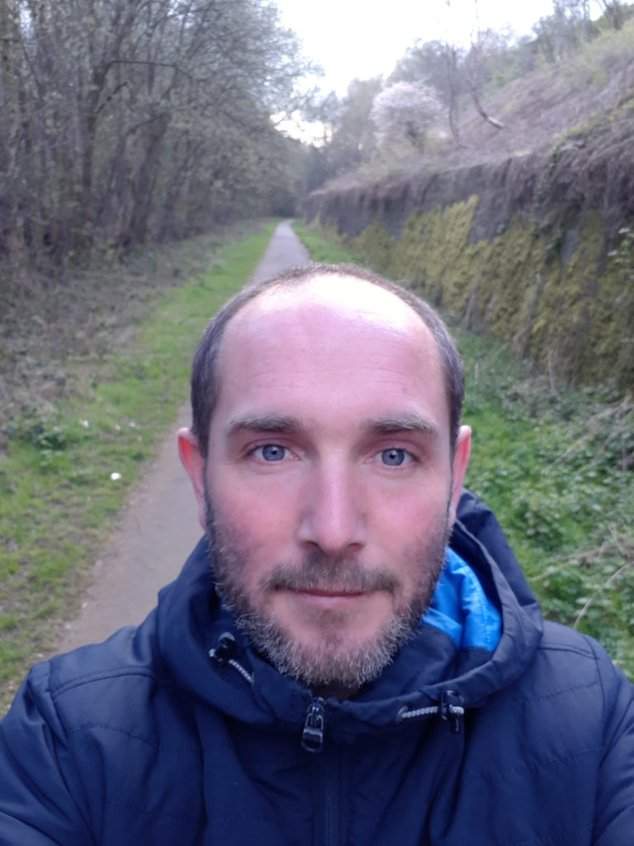 An image of Paul, a white man with greying stubble. He appears to be on a walk, there is a country lane framed by trees behind him. He is wearing a dark blue padded jacket.
