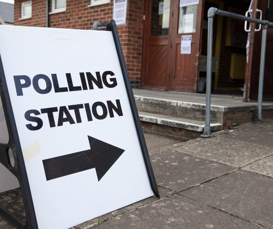 An image of a large white sign with black lettering which says, Polling Station. Underneath the lettering is a black arrow pointing to a wooden door which is open. There is a concrete ramp and also concrete stairs. 