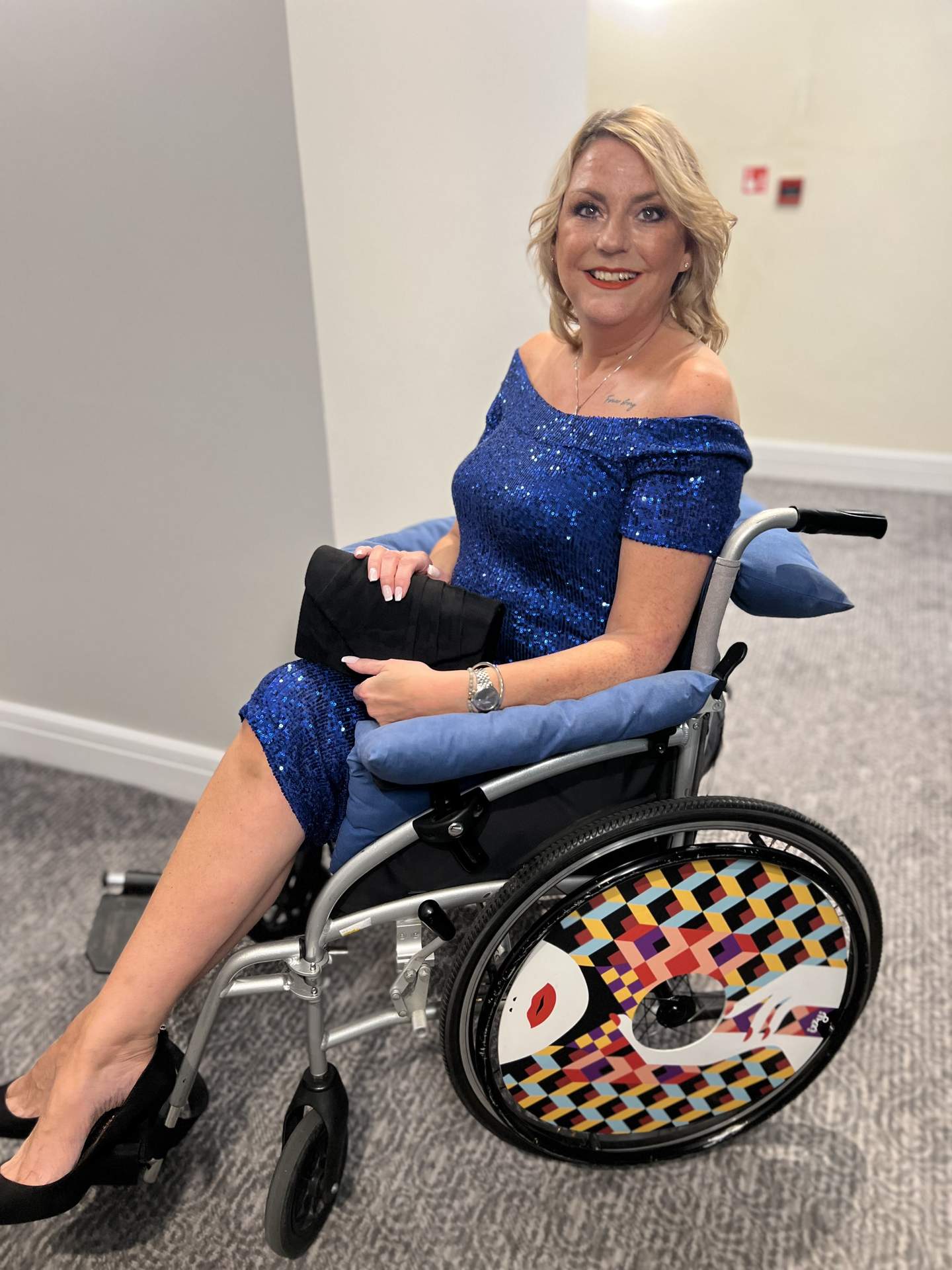 A picture of Jenn, who is a lady with blonde hair and is sat in a wheelchair wearing a sparkly blue dress. Her wheelchair has her designer spoke covers on from Izzy Wheels.