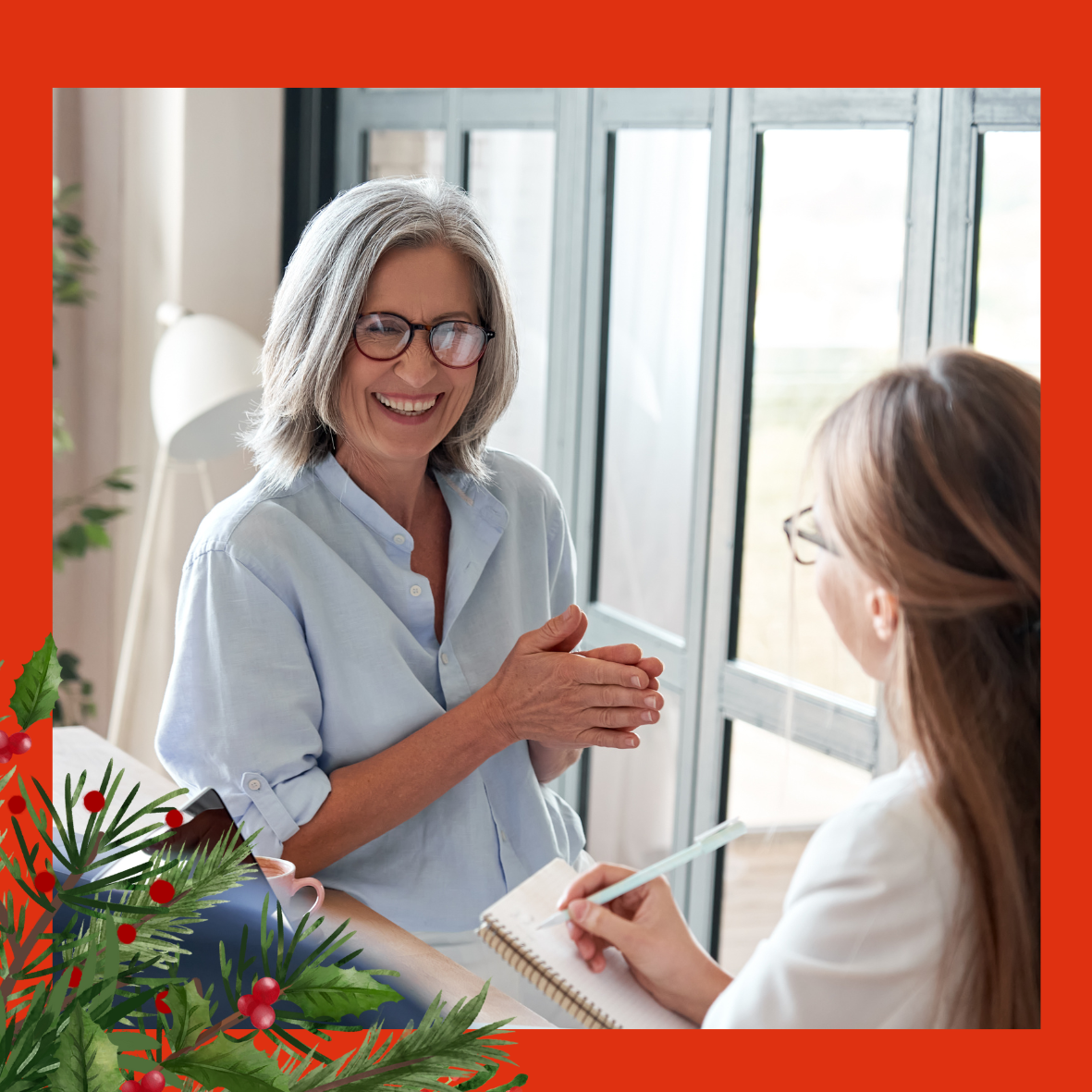 An image of a lady with red rimmed glasses, who is laughing with the woman opposite her. She has her hands together as if she is explaining something. The woman opposite is taking notes on a notepad. There are holly and red berries in the left hand corner of the image. 