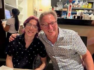 A picture of Jenn's son Mikey, who is a young man with dark purple coloured hair and is wearing glasses. He is sat next to Jenn's husband Matt who is a man in a patterned shirt and wearing glasses. Matt is smiling and has his arm around Mikey's shoulders.