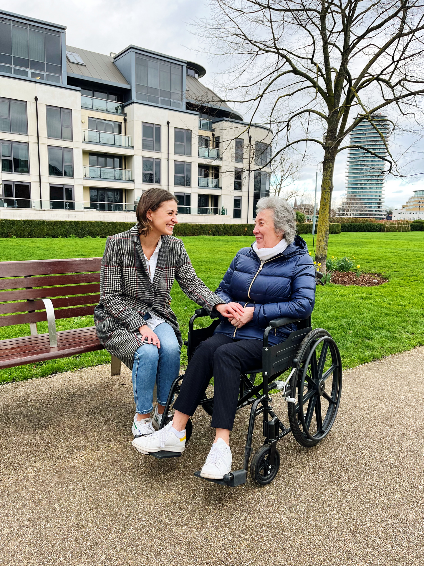 An an image of a woman in a checked coat and blue jeans, sat on a bench with an older woman in a wheelchair parked next to her. They are laughing and the younger woman is touching the older woman's hands.