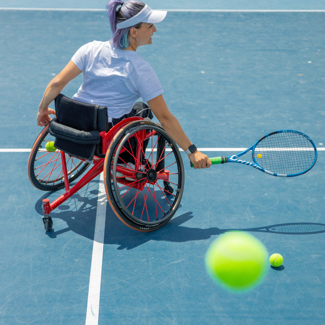 An image of a woman with purple and blue hair, wearing a white visor. She is sat in a red sports wheelchair and is holding a blue tennis racket.