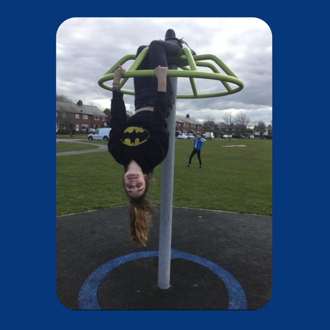 An image of Chloe hanging upside-down by her legs from a climbing frame at a park. She is grinning and wearing a batman hoodie. 