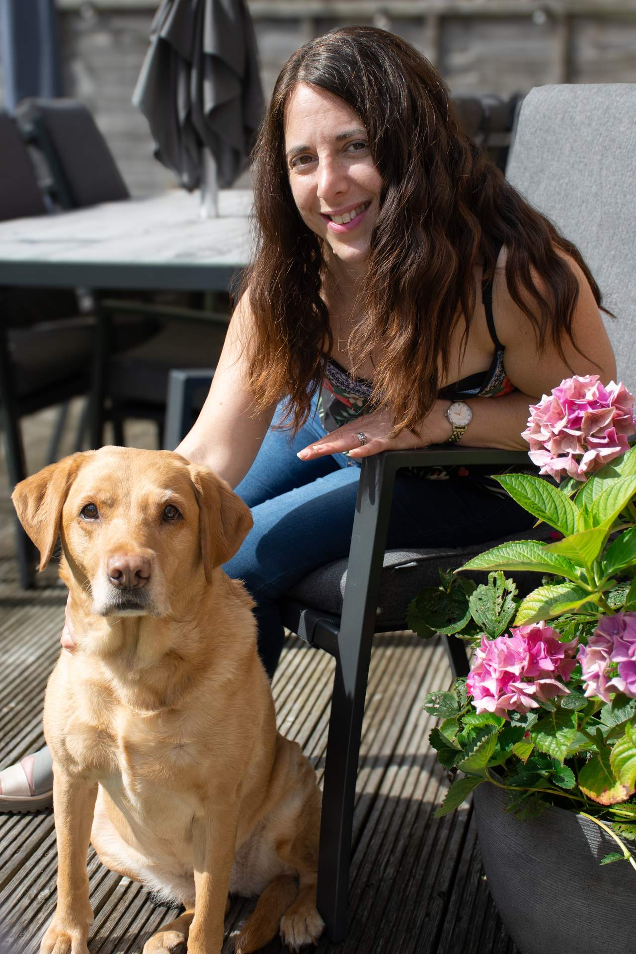 This image shows Georgina, who is a woman sitting outdoors on a patio, smiling warmly at the camera. She has long, wavy brown hair and is wearing a floral top and jeans. Beside her, sitting on the ground, is a golden-coloured Labrador dog, also looking directly at the camera. The setting includes grey outdoor furniture, with a table and chairs in the background. To the right, there is a plant pot with vibrant pink hydrangea flowers, adding a colourful touch to the scene. The overall mood is calm and cheerful, with natural sunlight illuminating the space.