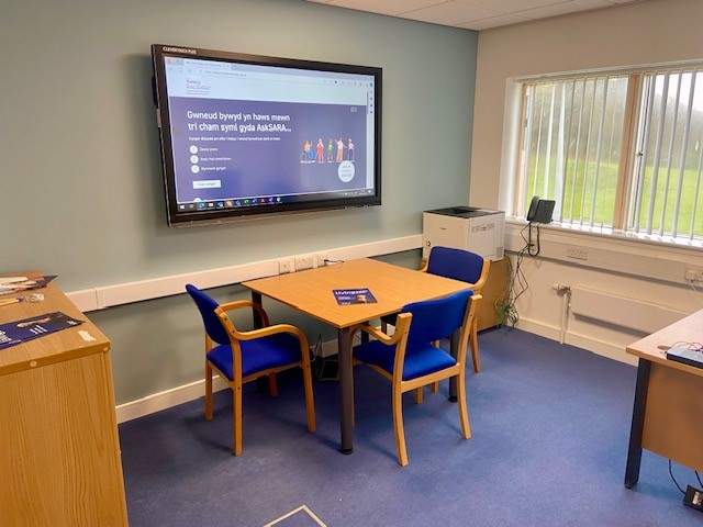 An image of a large screen on the wall demonstrating an AskSARA demonstration in the Welsh Language. In the room is a table with three chairs tucked in around it.