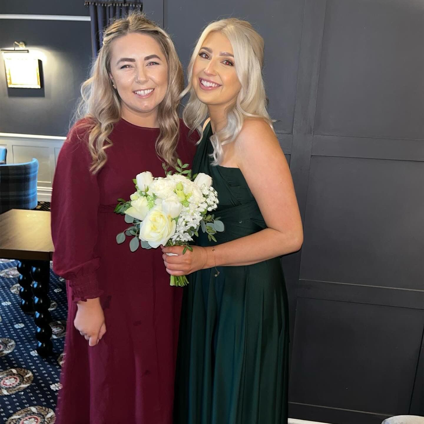 An image of Lucy and Johanna. Lucy is a woman wearing a maroon coloured dress, with blonde hair who is smiling. Next to her is Johanna, also with blonde hair and smiling. She is wearing a dark green occasion dress and is holding a bouquet of white flowers.