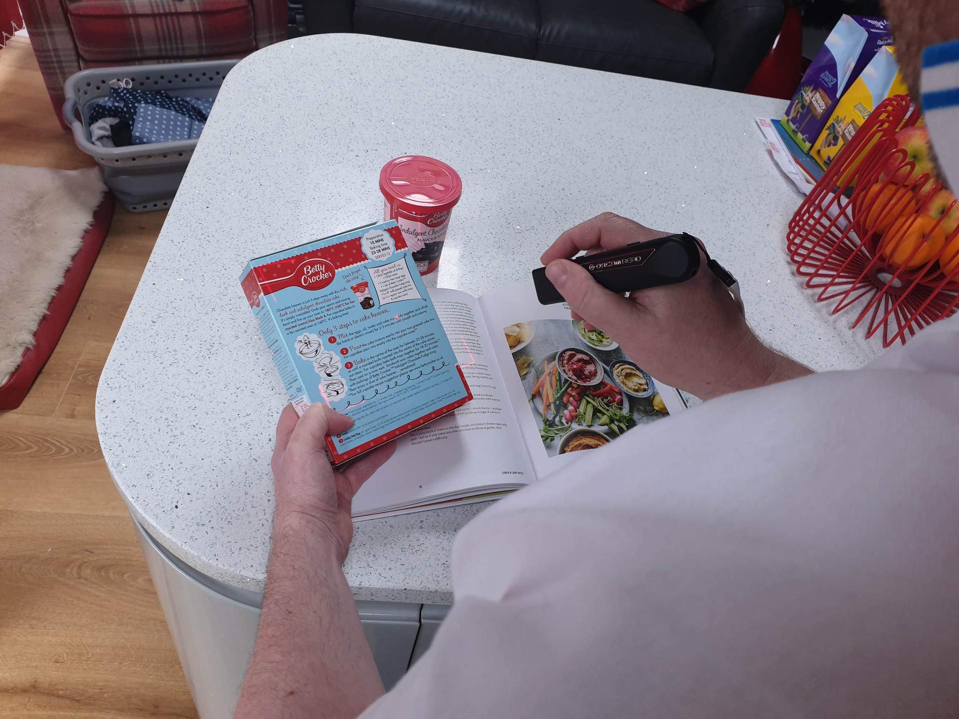 A person is holding the OrCam Read Smart handheld text to speech device, to capture information from a boxed cake mix.