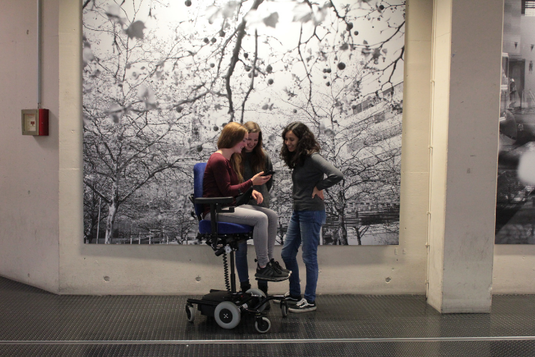 Three girls, one in a powered wheelchair, elevated, the others standing, in front of an art canvas. 