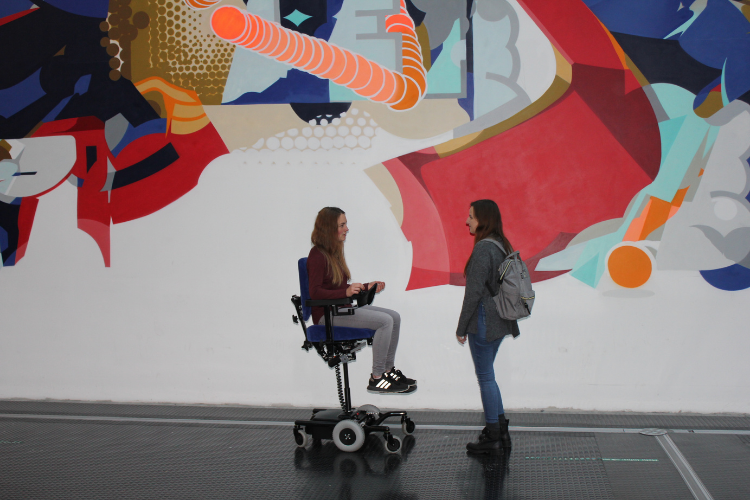 Two girls, one in a powered wheelchair, seat elevated, in front of a painted wall
